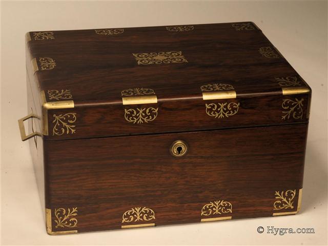 Figured rosewood box with rounded edges in alternating brass and rosewood having fine inlaid brass accents depicting  stylized neo-classical motifs . The box is of high quality, which continues into the compartmentalized interior. There is a lockable side drawer of dovetail construction fitted for jewelry. It is lined in velvet and embossed leather. The box has its original leather covered lift-out tray. There is a document wallet in the lid with a mirror behind.  Both the main lock and the drawer lock are working and have the same key. Circa 1825   Enlarge Picture